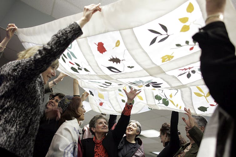 A handful of women smile as they hold up a white piece of fabric embroidered with colorful flowers.