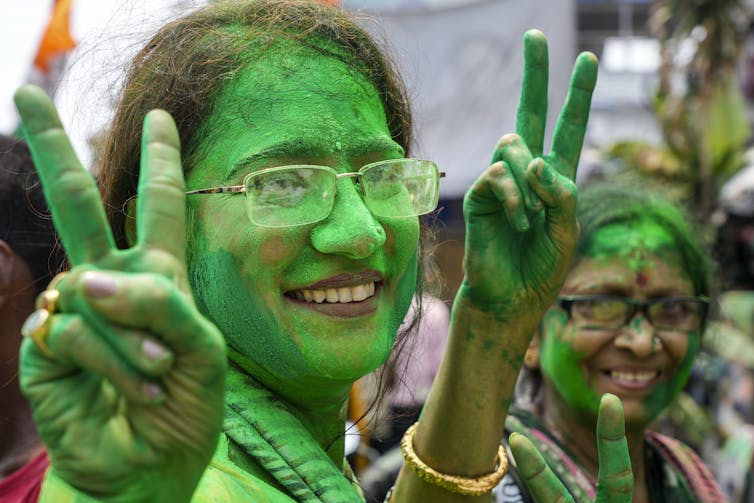 Faces of two women smeared with green color holding their fingers up in a victory sign.