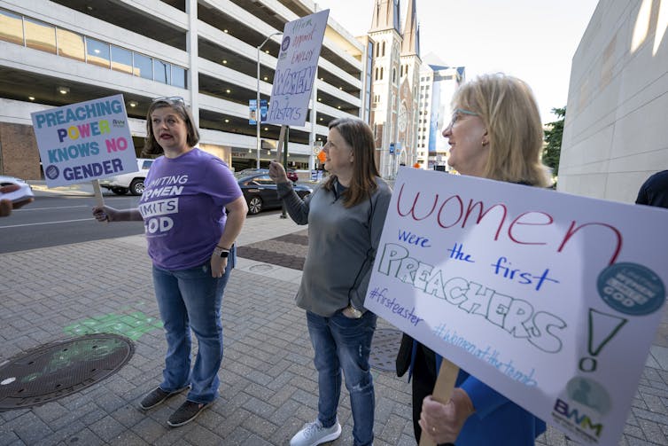 Three women standing together while holding placards. One of them reads, 'Preaching power knows no gender.'