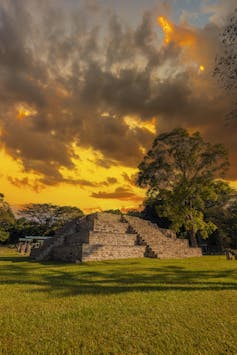 A brick-colored structure with a staircase on each side sits in a grassy area among trees at sunset.