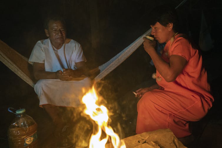 Two men sit around a large fire with one drinking from a glass.