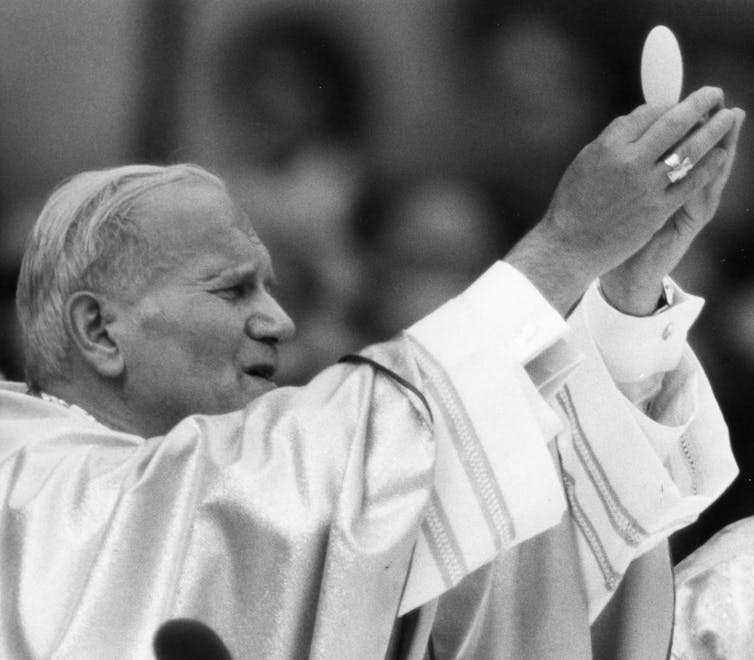 An elderly man in a white gown and cap holds up his hands as though in a prayer.